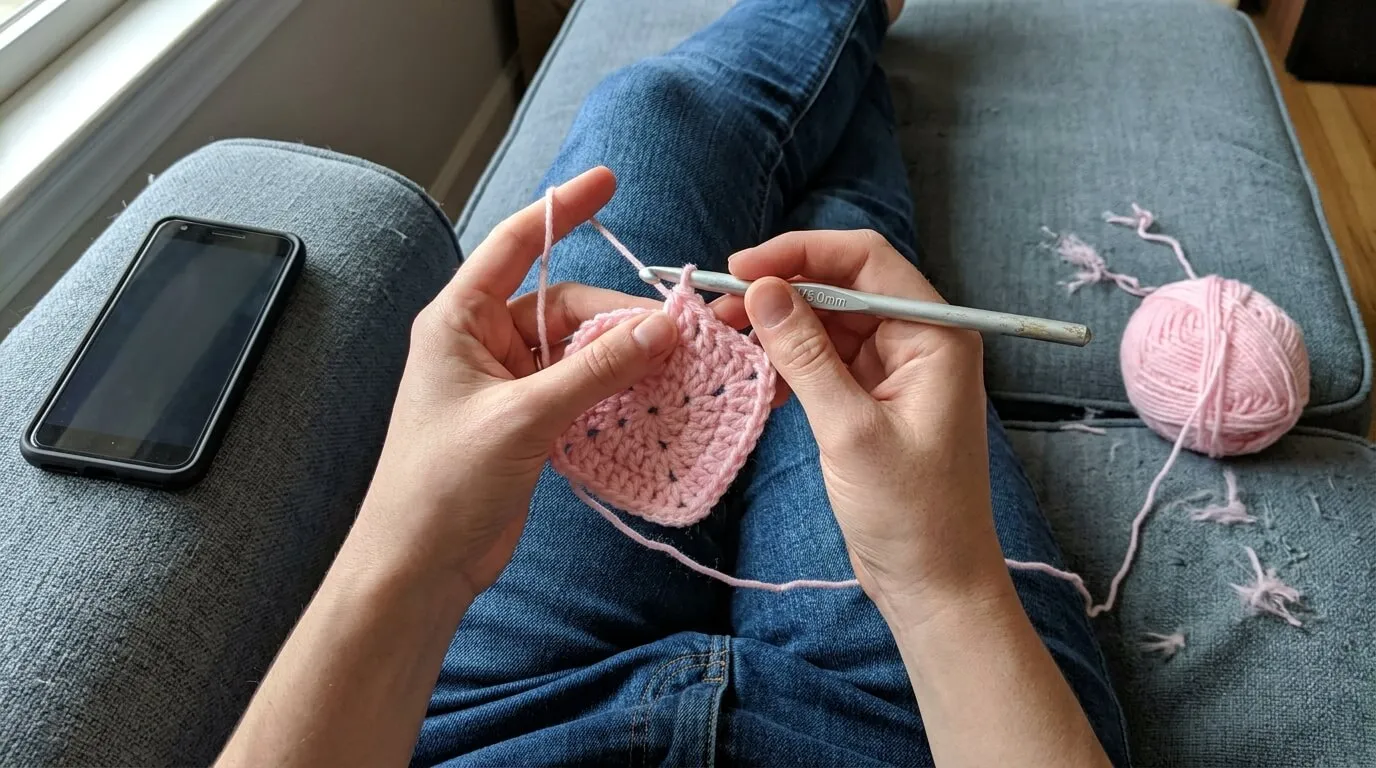 Mãos fazendo crochê com fio colorido no sofá de casa, com o celular apoiado no braço do sofá